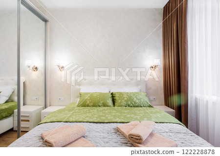 A well-appointed small modern bedroom with a cozy design, featuring a queen-sized bed, bedside tables. A large window with curtains allowing soft daylight. 122228878