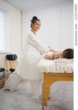 Massage process. Beautiful smiling woman in white outfit makes body massage to lying client in pro salon, vertical image, selective focus. 122229982