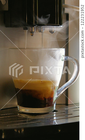 A close-up of a coffee machine pouring a cappuccino into a clear glass cup. The coffee is layered with espresso, milk foam, and a layer of steamed milk. Steam rises from the cup and the machine. A close-up of a coffee machine pouring a cappuccino into a clear glass cup. The coffee is layered with espresso, milk foam, and a layer of steamed milk. Steam rises from the cup and the machine. 122231952