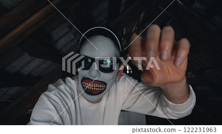 A scary masked man in a long-sleeved white sweatshirt and black cap stretches out his hand in a dimly lit attic with wooden beams. A scary masked man in a long-sleeved white sweatshirt and black cap stretches out his hand in a dimly lit attic with wooden beams. 122231963