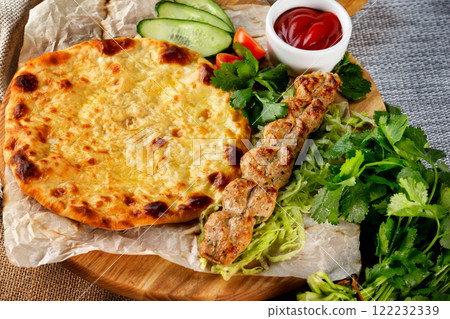 Traditional georgian food - shish kebab and khachapuri pie served with fresh vegetables at wooden tray board on table with sack cloth background. Traditional georgian food - shish kebab and khachapuri pie served with fresh vegetables at wooden tray board on table with sack cloth background. 122232339
