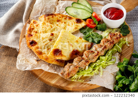 Traditional georgian food - shish kebab and khachapuri pie served with fresh vegetables at wooden tray board on table with sack cloth background. Traditional georgian food - shish kebab and khachapuri pie served with fresh vegetables at wooden tray board on table with sack cloth background. 122232340