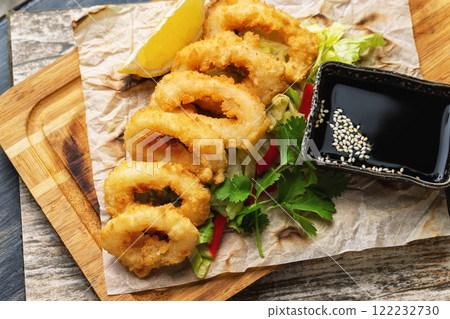 Top view image of golden crispy dried calamari rings with soy sauce served on paper at wooden table background. Top view image of golden crispy dried calamari rings with soy sauce served on paper at wooden table background. 122232730