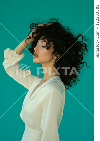 Graceful woman with curly hair in ivory dress against teal. 122233585