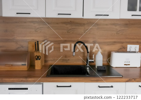 A black kitchen sink with a black flexible faucet, it has dishwashing detergent in a white bottle and a black dishwashing sponge against the background of a modern kitchen with white cabinets and a A black kitchen sink with a black flexible faucet, it has dishwashing detergent in a white bottle and a black dishwashing sponge against the background of a modern kitchen with white cabinets and a 122233717