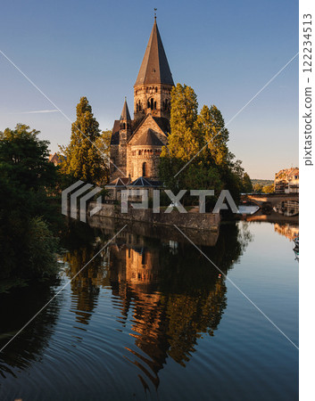 Historic stone church with tall spires surrounded by trees, reflected in a calm river under a soft golden evening light Historic stone church with tall spires surrounded by trees, reflected in a calm river under a soft golden evening light 122234513