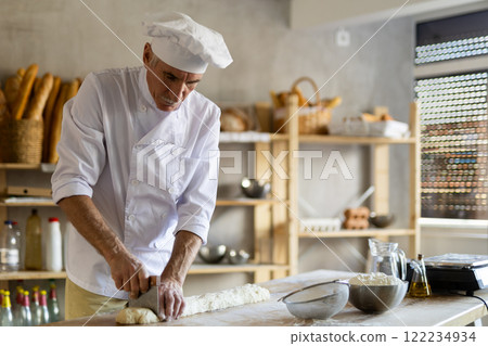 Elderly experienced male baker using knife to cut raw dough to make croissants 122234934