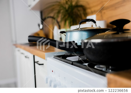 Close-up of a black frying pan with a transparent lid and a silicone handle and a blue saucepan with a transparent lid and silicone handles against the background of a modern wooden-white kitchen out 122236656