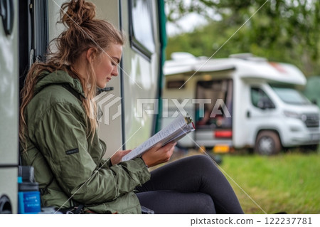 A young woman enjoys nature while reading outside her camper. Surrounded by lush greenery, she finds inspiration in the peaceful setting. Generative AI. 122237781