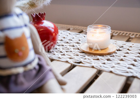 lit candle sits on macrame woven runner and soft doll on wooden table. warmth to the cozy indoor 122239303