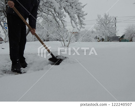 A man in black clears the snow-white crust with a shovel in a village A man in black clears the snow-white crust with a shovel in a village 122240507