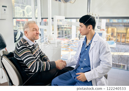 Dentist, smiling warmly, shakes hand of senior man, patient reclined in chair, concluding consultation in modern, sunlit dental care office. Dentist, smiling warmly, shakes hand of senior man, patient reclined in chair, concluding consultation in modern, sunlit dental care office. 122240533