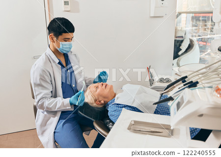 Male doctor examining elderly woman in dental chair, using tools, calm atmosphere in organized clinic with bright natural lighting. 122240555