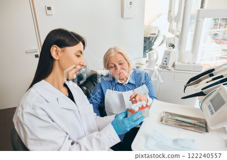 Female doctor demonstrating dental model to elderly woman, seated in well-equipped dental clinic, both engaged in conversation. Female doctor demonstrating dental model to elderly woman, seated in well-equipped dental clinic, both engaged in conversation. 122240557