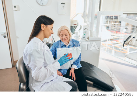 Female doctor in gloves and mask explaining dental model to elderly woman seated beside dental chair in well-lit clinic. Hygiene. 122240593