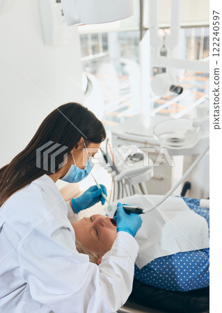 Doctor leaning over elderly woman during dental examination, using special tools, in clean and bright clinic setting with visible dental equipment. 122240597