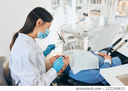 Elderly woman in dental chair being examined by female doctor in white coat, gloves, and mask, using dental tools, bright modern office environment. 122240615