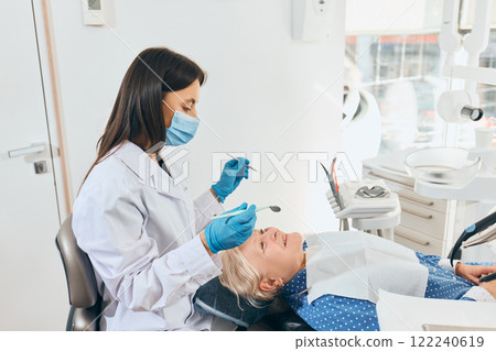 Female doctor in mask and gloves examining mouth of elderly woman lying in dental chair, using dental tools, equipment visible in bright dental office. Female doctor in mask and gloves examining mouth of elderly woman lying in dental chair, using dental tools, equipment visible in bright dental office. 122240619