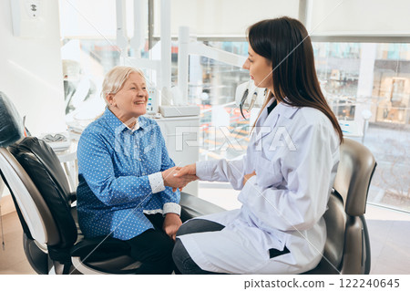 Elderly woman seated in dental chair shaking hands with female doctor in lab coat, smiling, medical tools and equipment visible in bright room. 122240645