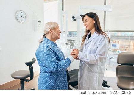 Female doctor warmly holding hands with elderly woman in blue polka-dot shirt, both smiling, standing in well-lit medical office 122240650