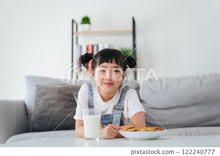 A young girl is sitting on a couch with a glass of milk and a plate of cookies. 122240777