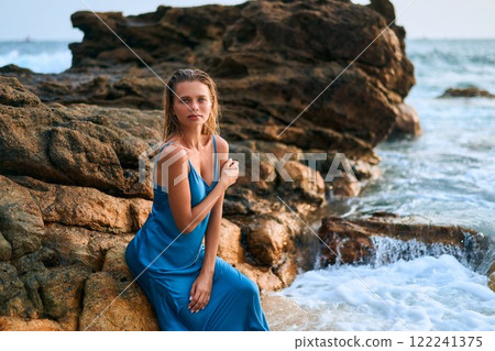Elegant woman in wet dress sits by ocean rocks on tropical island. Waves crash. Model poses by sea showcasing beauty and nature contrast. Exotic travel destination with serene coastal vibe. Elegant woman in wet dress sits by ocean rocks on tropical island. Waves crash. Model poses by sea showcasing beauty and nature contrast. Exotic travel destination with serene coastal vibe. 122241375