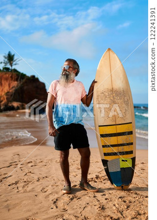 Senior man with surfboard stands on sandy beach. Elderly surfer enjoys sunny day by ocean. Active lifestyle for older adults. Sea waves, blue sky in holiday setting. 122241391