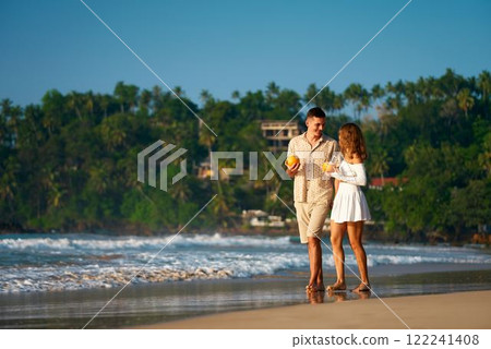Romantic stroll along sunlit tropical beach, couple walks barefoot near sea waves, intimate honeymoon moments, relaxed ambiance, love, travel theme with palms in background, sunset creates warm mood. 122241408