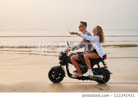 Young couple ride on motorbike. Man drives motorcycle on sandy beach. Woman points, enjoys adventure. Travel holiday vibes. Ocean waves, sunset horizon. Romantic journey, explore tropical shore. 122241431