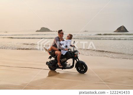 Romantic couple rides motorbike on beach at sunset. Man drives scooter, woman sits behind hugging him. Joyful lovers on honeymoon, explore seaside adventure. Tropical ocean background, freedom travel. Romantic couple rides motorbike on beach at sunset. Man drives scooter, woman sits behind hugging him. Joyful lovers on honeymoon, explore seaside adventure. Tropical ocean background, freedom travel. 122241433