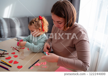Caucasian adult mother and her little cute daughter cut out paper hearts sitting at a desk. Family is preparing for the celebration of Valentine's Day Caucasian adult mother and her little cute daughter cut out paper hearts sitting at a desk. Family is preparing for the celebration of Valentine's Day 122244800