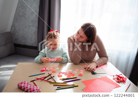 Caucasian woman kindergarten teacher teaches a little happy girl how to cut hearts out of red paper for Valentine's Day. Wide shot 122244801
