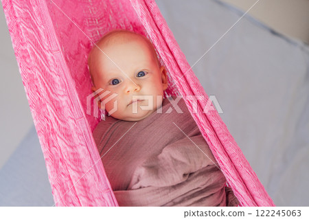 A 3-month-old baby peacefully falls asleep in a soft muslin swaddle while cradled in a cozy hammock. A serene and tender moment of comfort and relaxation. Baby care, rest, and tranquility concept A 3-month-old baby peacefully falls asleep in a soft muslin swaddle while cradled in a cozy hammock. A serene and tender moment of comfort and relaxation. Baby care, rest, and tranquility concept 122245063