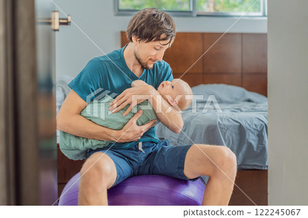 A father sits on a fitness ball in a cozy bedroom, gently rocking his baby wrapped in a soft muslin swaddle. A nurturing and soothing moment of fatherhood and care. Parenting, bonding, and comfort A father sits on a fitness ball in a cozy bedroom, gently rocking his baby wrapped in a soft muslin swaddle. A nurturing and soothing moment of fatherhood and care. Parenting, bonding, and comfort 122245067