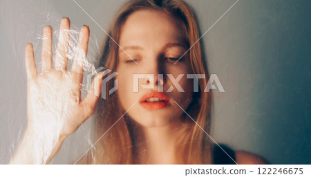 Female isolation. Sadness art. Sensual disturbed woman standing behind wrinkled polyethylene film on blur pastel pink blue color gradient glow background. Female isolation. Sadness art. Sensual disturbed woman standing behind wrinkled polyethylene film on blur pastel pink blue color gradient glow background. 122246675