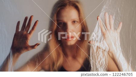 Female sorrow. Freedom desire. Disturbed woman trapped behind wrinkled transparent polyethylene film on gray out of focus background. Female sorrow. Freedom desire. Disturbed woman trapped behind wrinkled transparent polyethylene film on gray out of focus background. 122246705