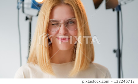 Female posing. Positive photo session. Confident blonde girl smiling winking at video camera. Cheerful woman closeup glasses spotlighted pro studio. 122246712