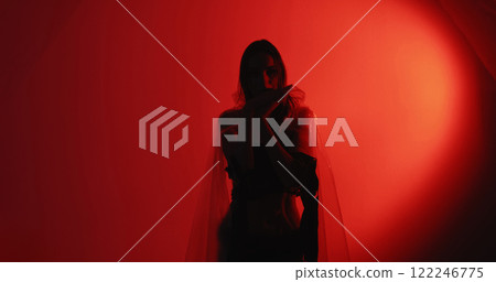 Dance therapy. Calming practice. Peaceful woman silhouette in cellophane rags performing hypnotic hand movements isolated on dark red glowing copy space background. Dance therapy. Calming practice. Peaceful woman silhouette in cellophane rags performing hypnotic hand movements isolated on dark red glowing copy space background. 122246775