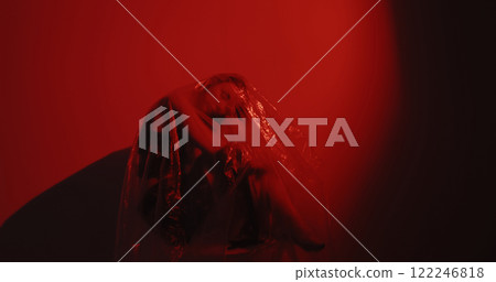 Woman protection. Sleeping beauty. Depressed tired hurt female sitting covered with transparent polyethylene film on dark red flicker glow out of focus background. 122246818