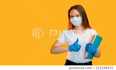 Like gesture. Socially distanced. Confident female teacher in protective face mask gloves with books showing thumb up isolated on yellow copy space background. 122246932