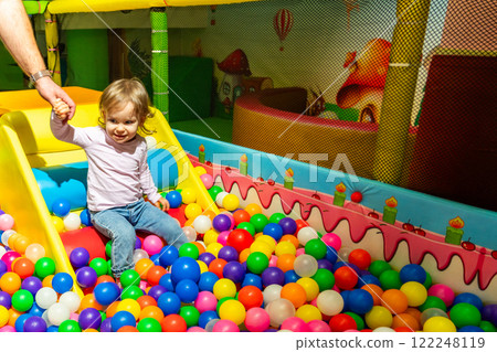 A joyful toddler sliding into a vibrant ball pit under adult supervision. 122248119