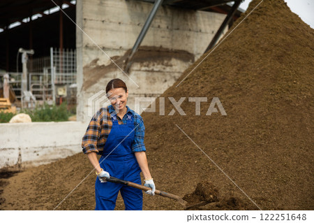 Female worker in blue jumpsuit stands next to pile of manure. Female worker in blue jumpsuit stands next to pile of manure. 122251648