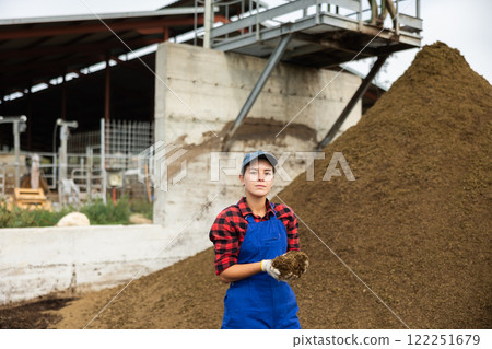 Female farmer with shovel poses against pile of dried and crushed cow dung in the backyard of farm 122251679