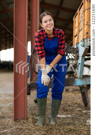 Portrait of young girl working in cowshed in blue overalls and boots 122251680