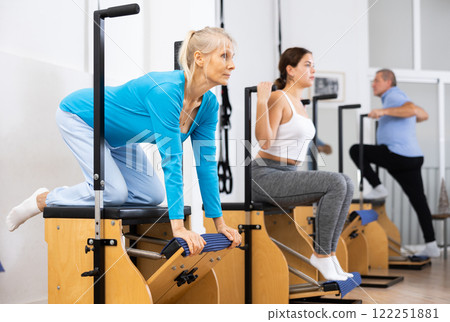 Mature European woman making Pilates exercising while standing on knees using Wanda chair machine at gym Mature European woman making Pilates exercising while standing on knees using Wanda chair machine at gym 122251881