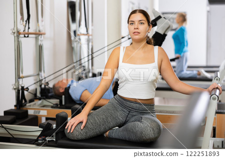 Positive young female performing mermaid exercise on reformer Pilates machine in studio Positive young female performing mermaid exercise on reformer Pilates machine in studio 122251893