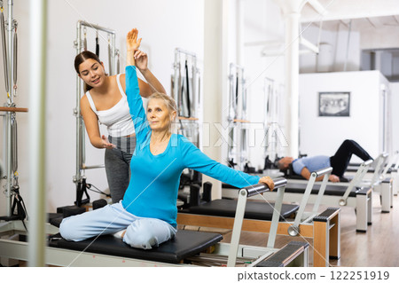 Smiling personal female trainer assisting senior woman exercising on Pilates machine in rehabilitation center 122251919