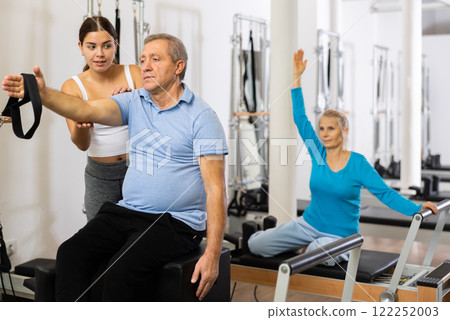 Positive young female instructor controlling movements of elderly man stretching on Pilates reformer equipment in studio Positive young female instructor controlling movements of elderly man stretching on Pilates reformer equipment in studio 122252003