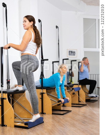 Relaxed young female standing and exercising with Pilates Wanda chair during group training in rehabilitation center Relaxed young female standing and exercising with Pilates Wanda chair during group training in rehabilitation center 122252010