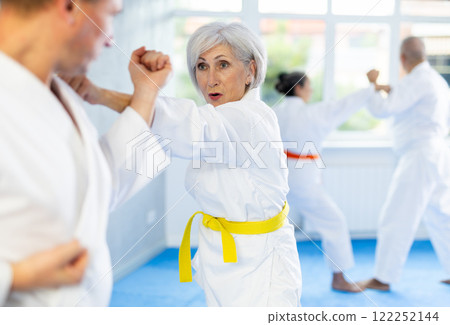 Group of mature people in kimono sparring together in gym during karate training Group of mature people in kimono sparring together in gym during karate training 122252144